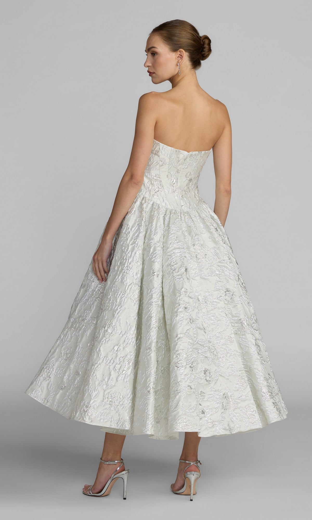 A woman in the Mac Duggal 69146 tea-length party dress, featuring a textured white brocade fabric and a strapless sweetheart bodice, stands with her back to the camera, looking over her shoulder while wearing silver heels against a plain gray background.